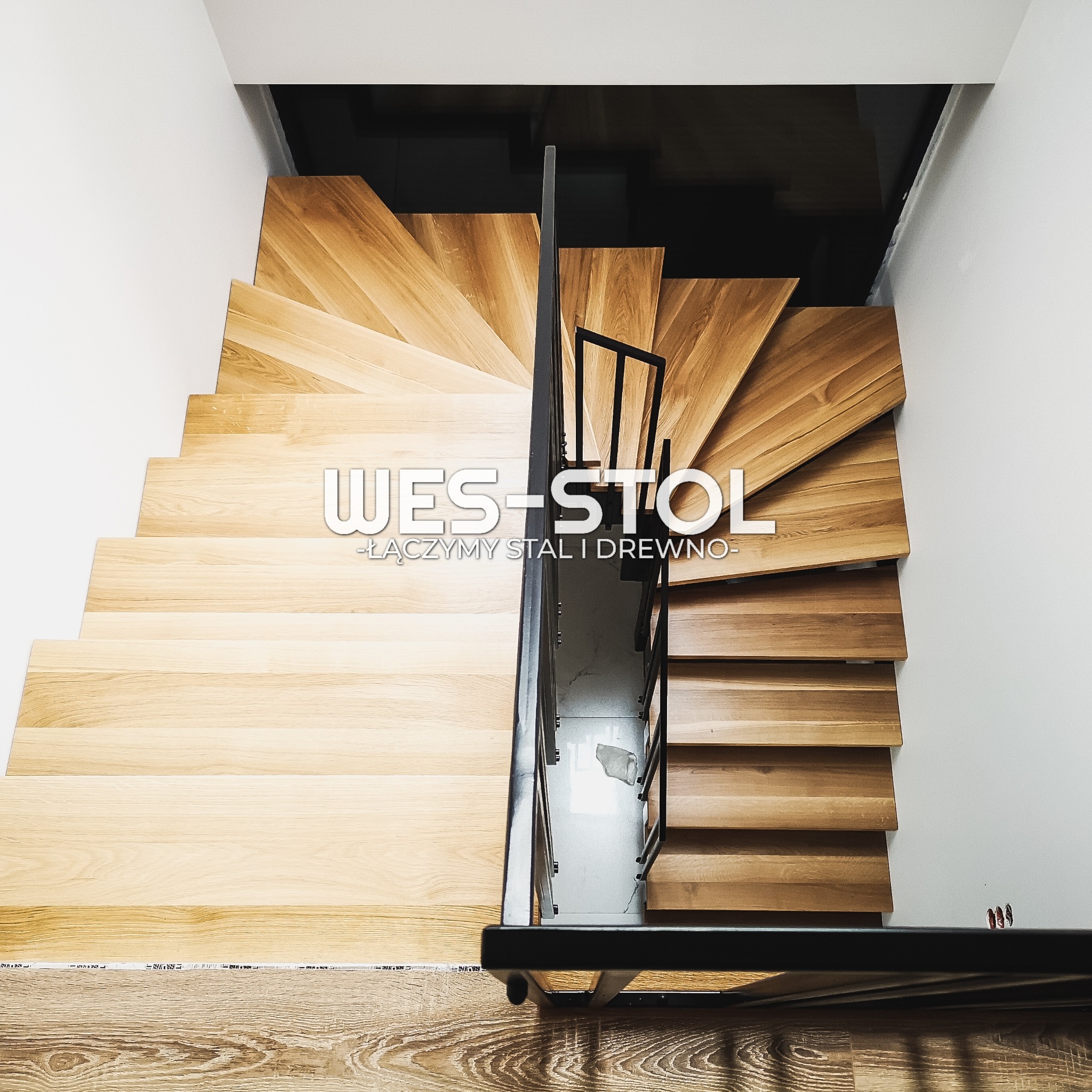 Spiralne schody z drewnianymi stopniami i metalową balustradą, widok z góry, logo firmy nałożone na obraz.