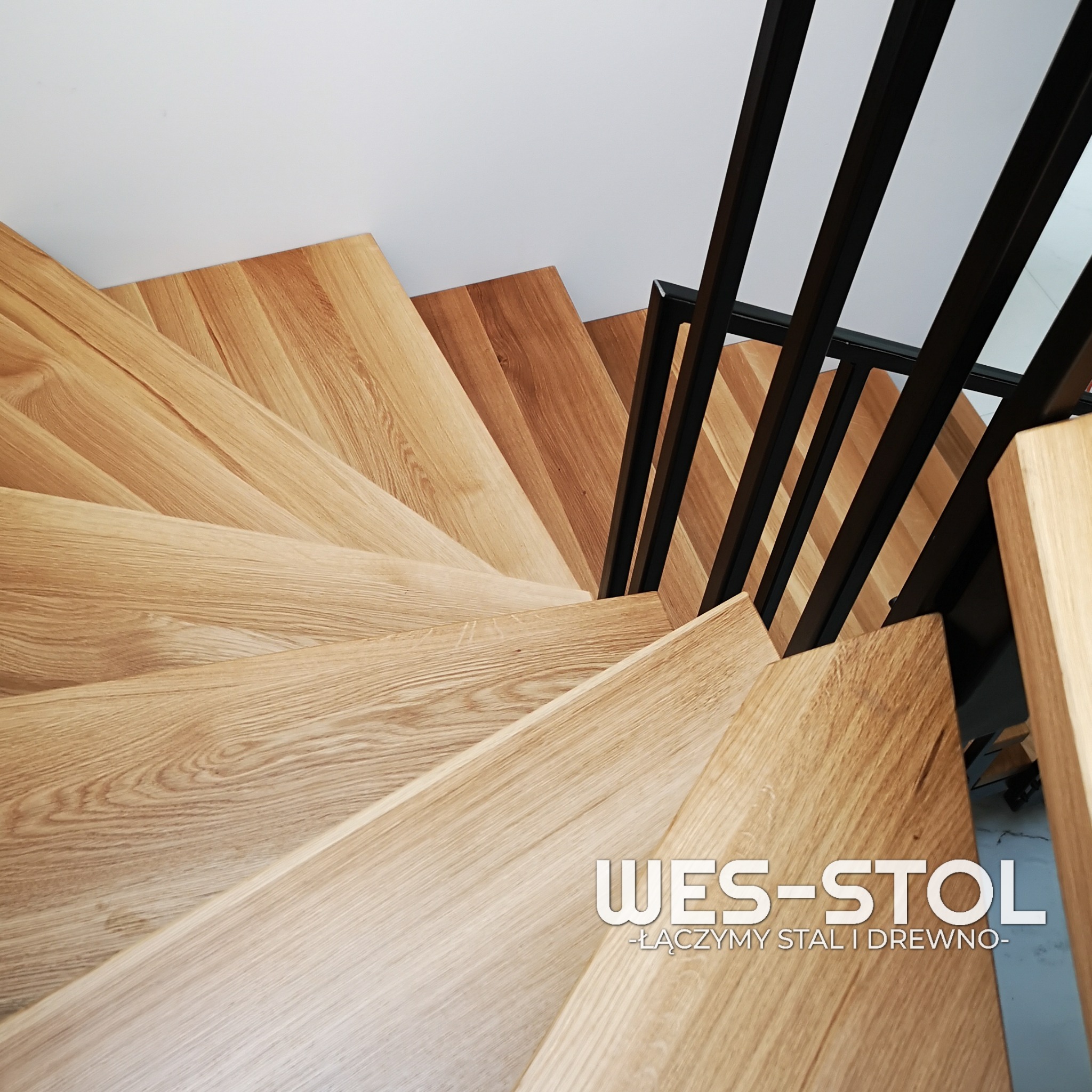 Widok z góry na kręte schody z jasnego drewna z czarną metalową balustradą. Na stopniach widoczne logo firmy WES-STOL.
