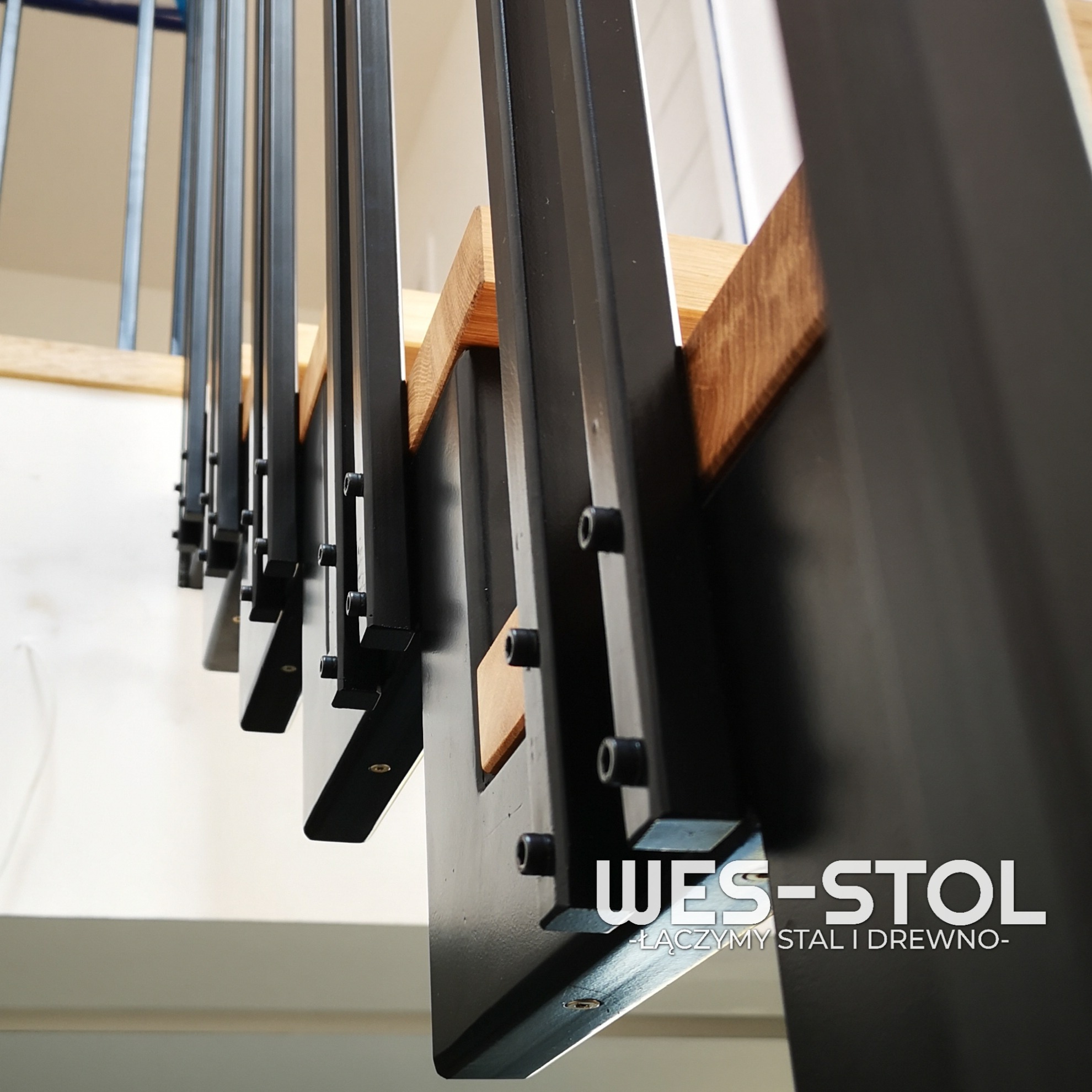 Metalowa konstrukcja schodów z drewnianymi stopniami, widoczne śruby mocujące, ujęcie z dołu, logo firmy na pierwszym planie.