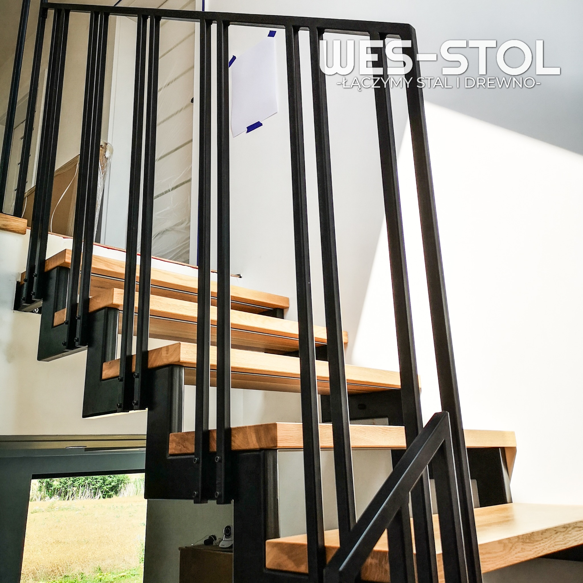 Schody metalowo-drewniane w stylu loft z czarną metalową balustradą i drewnianymi stopniami, widok od dołu, z naklejoną kartką na ścianie.