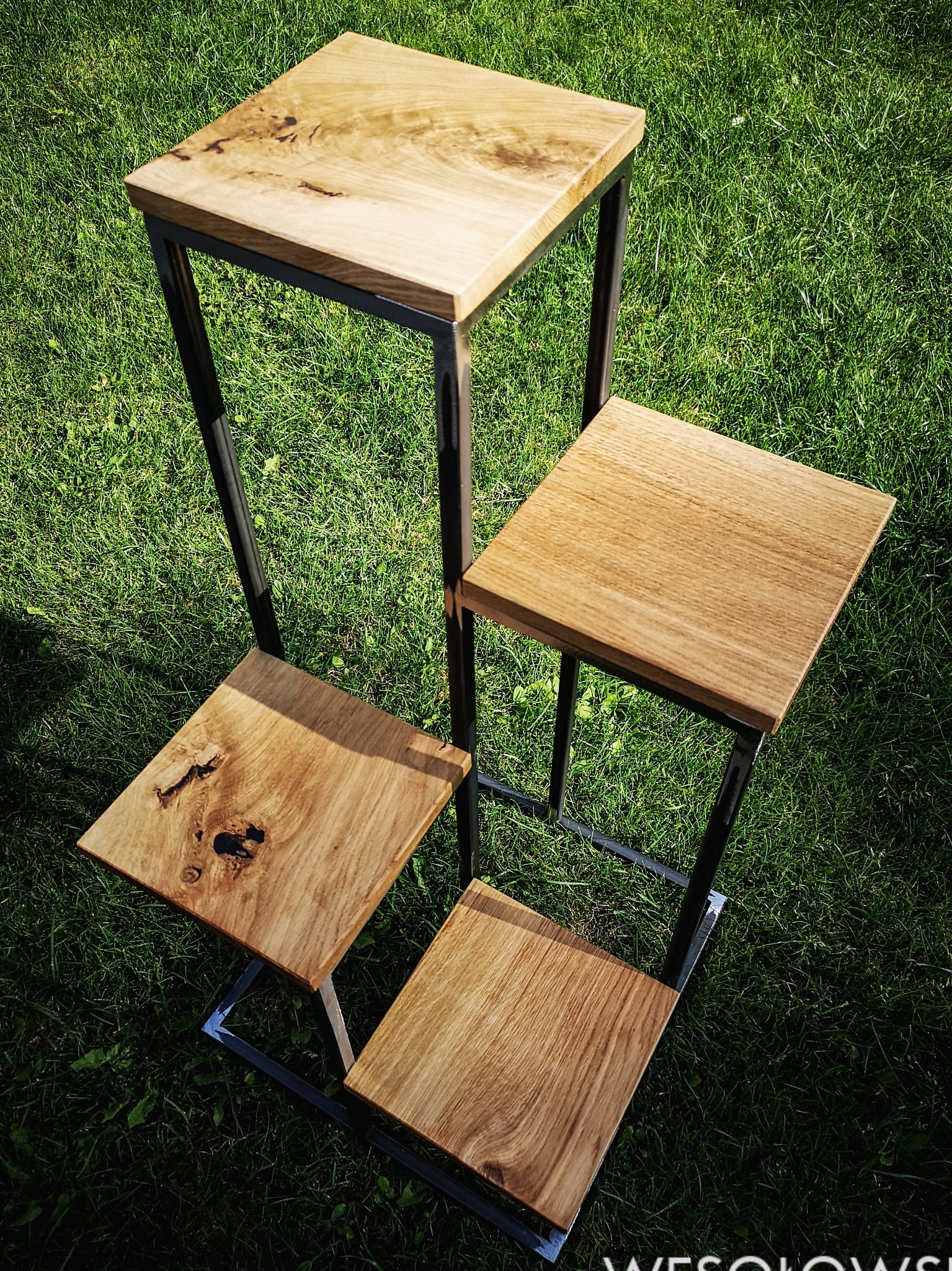 Czteropoziomowy kwietnik w stylu loft z drewnianymi blatami o zróżnicowanej strukturze i metalowym stelażem, ustawiony na trawie.