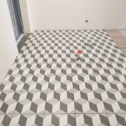 Podłoga z płytek w geometryczny wzór 3D w trakcie układania, widoczne krzyżyki dystansowe, śrubokręt i okulary na płytkach, fragment nieukończonej podłogi z drewnopodobnych paneli.