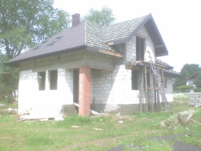 Dom w trakcie budowy z częściowo położonym brązowym dachem z blachy trapezowej i widoczną więźbą dachową oraz rusztowaniem z drabiny.