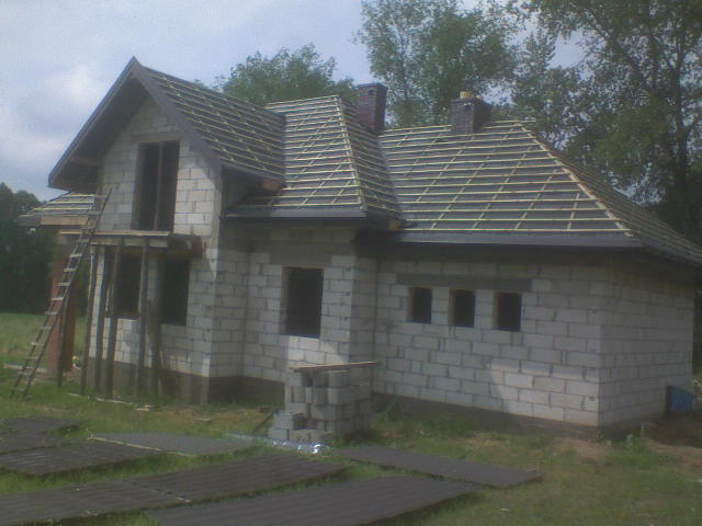 Niedokończony dom z widoczną więźbą dachową i przygotowanymi płytami dachowymi na trawie, widoczny drabina oparta o ścianę.