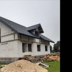 Surowy stan domu jednorodzinnego z czarną dachówką, widoczny stos drewna i piasku przed budynkiem.