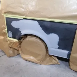 Przygotowanie nadkola samochodu do lakierowania, zabezpieczone papierem maskującym i taśmą, widoczne szpachlowanie i podkład.