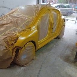 Auto Naprawa - Żółty samochód w warsztacie, przygotowany do lakierowania, zakryty papierem i folią. Widoczne otwarte drzwi i fragment nadkola.