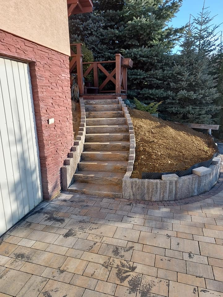 Ułożona kostka brukowa prowadząca do schodów z obrzeżami, na których znajduje się nasyp ziemi z roślinami, widok na budynek z garażem i drzewa iglaste w tle.