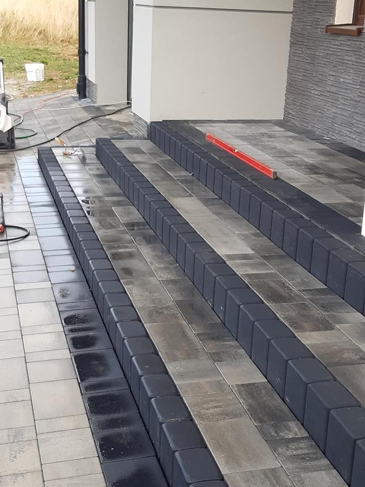 Schody wejściowe wyłożone kostką granitową w odcieniach szarości, obrzeża z ciemnej kostki, poziomica na jednym ze stopni, widoczny fragment elewacji budynku.