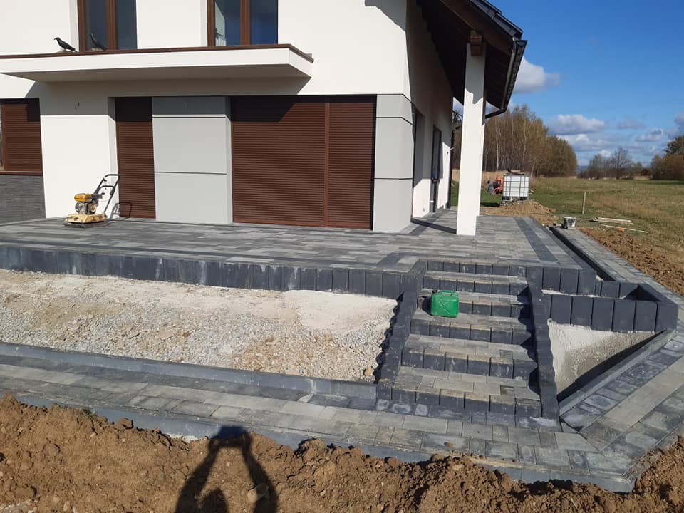 Nowo ułożona, szara kostka brukowa przed domem z widocznymi schodami i krawężnikami, zagęszczarka w tle, teren częściowo przygotowany pod dalsze prace.