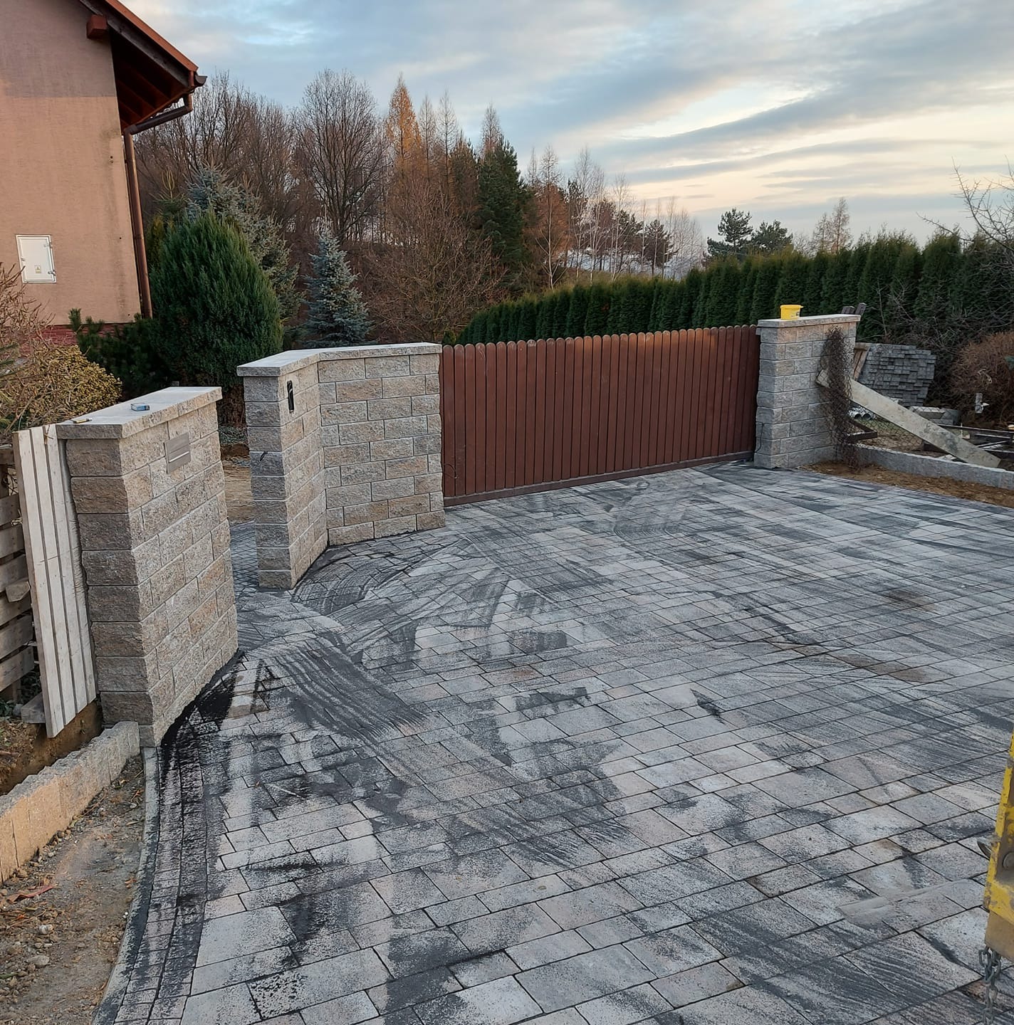Nowo ułożona szara kostka brukowa prowadząca do brązowej, drewnianej bramy wjazdowej z kamiennymi słupkami, widok na dom i drzewa w tle.
