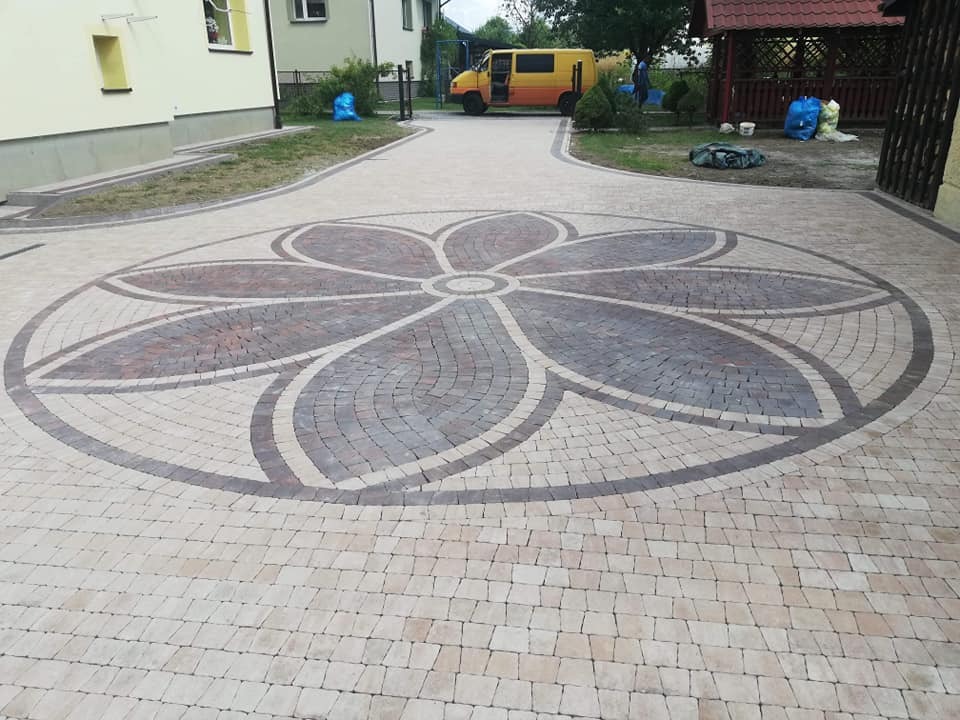 Ułożona kostka granitowa w kształcie kwiatu na podjeździe, z żółtym samochodem dostawczym i budynkiem w tle.