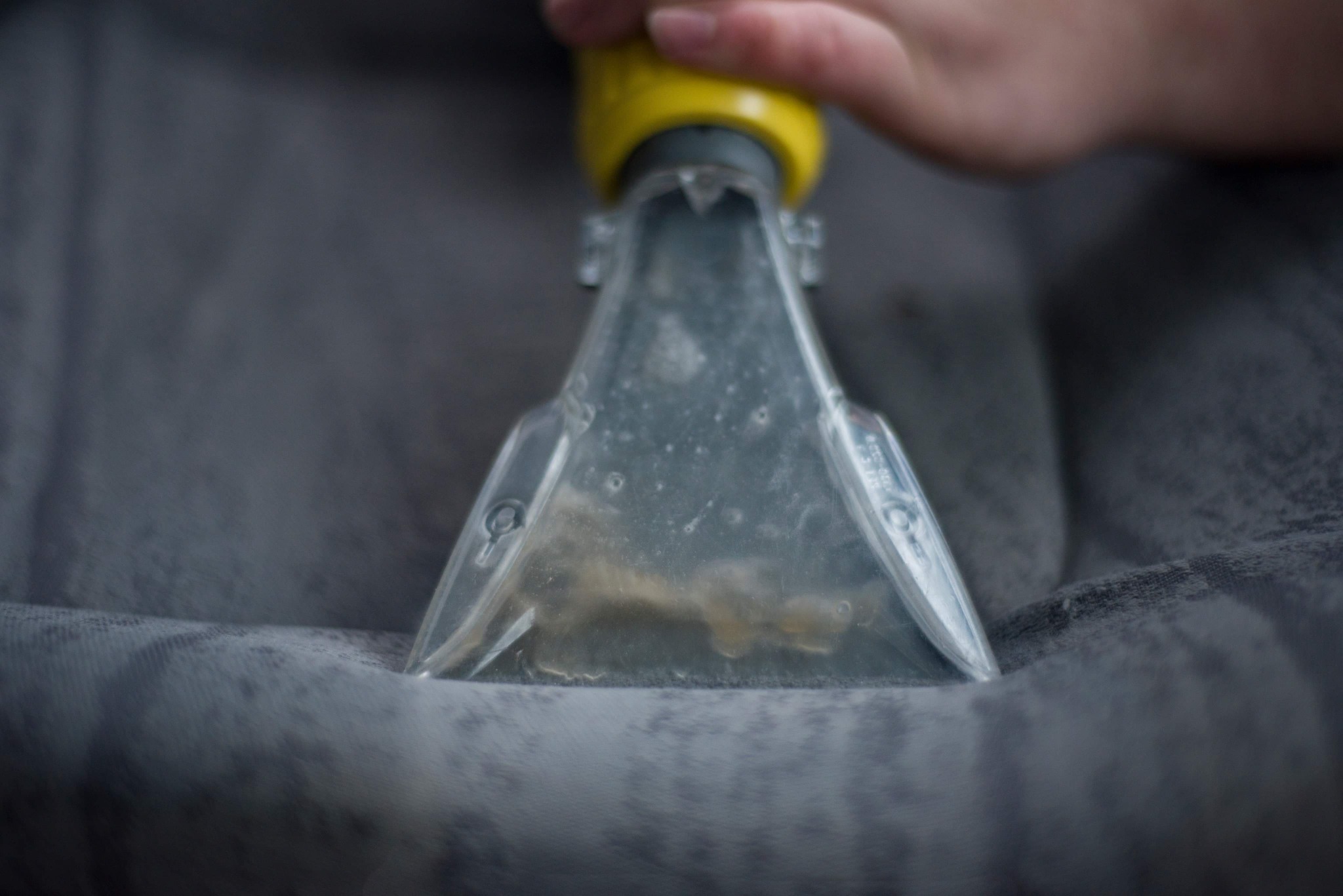 Dysza odkurzacza piorącego z żółtą obudową w trakcie czyszczenia szarej tapicerki, widoczny brud w zbiorniku.