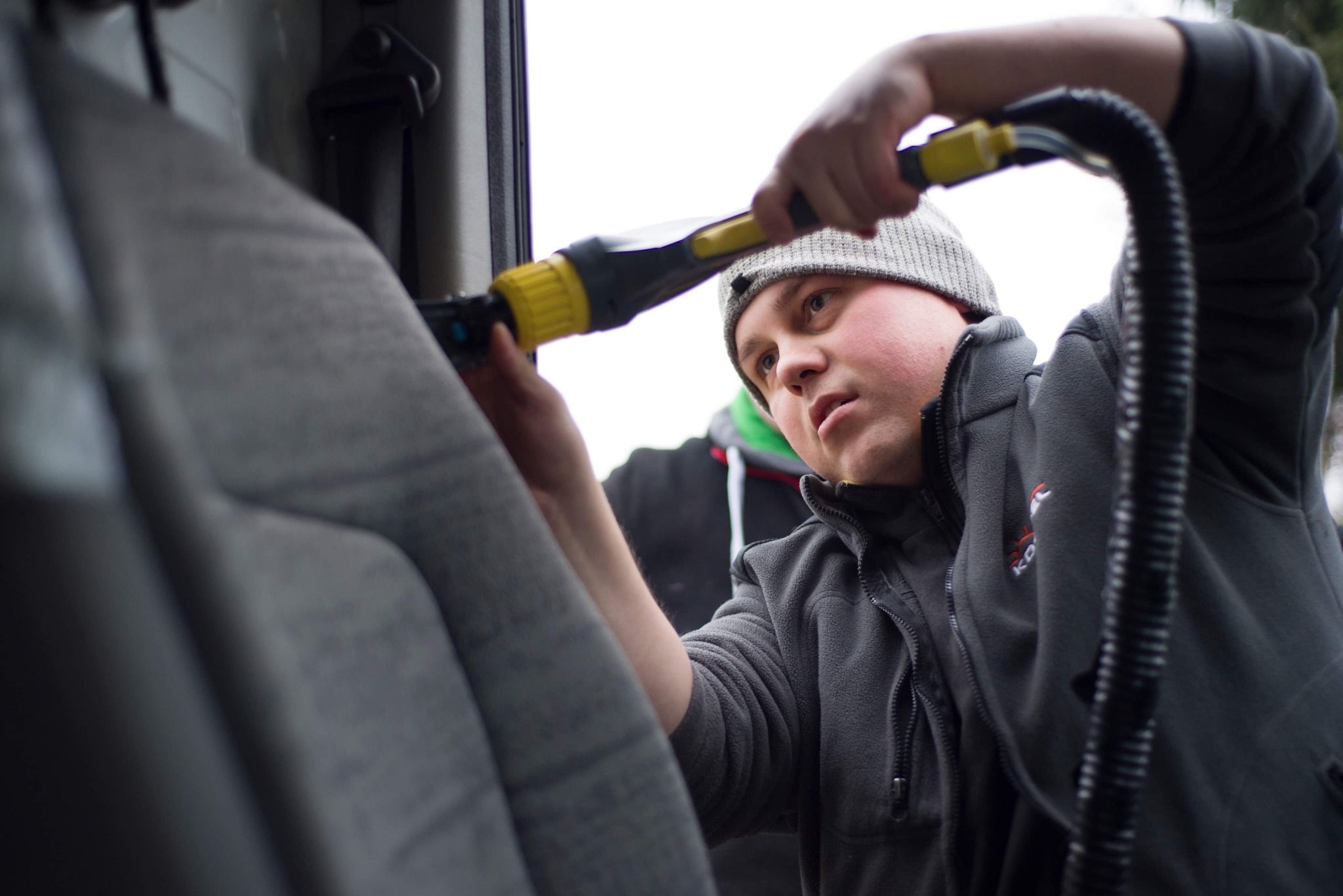 Mężczyzna w szarej kurtce i czapce czyści szary fotel samochodowy za pomocą odkurzacza z żółtą końcówką.