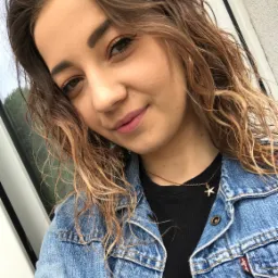 Portret uśmiechniętej kobiety w dżinsowej kurtce z zawieszką w kształcie gwiazdy na złotym łańcuszku, wykonany w pochmurny dzień na tle okna z widocznymi drzewami.