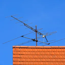 Antena telewizyjna zamontowana na dachu pokrytym czerwoną dach&oacute;wką, widoczna na tle błękitnego nieba.