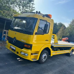 Żółta laweta Mercedes Atego z pomarańczowym światłem ostrzegawczym, zaparkowana na asfaltowym placu, z widocznym numerem rejestracyjnym i elementami wyposażenia.