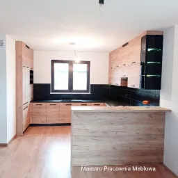 Jasna kuchnia z drewnianymi frontami i czarnym blatem, wyspa kuchenna z blatem o podobnym odcieniu, oświetlenie punktowe, widok na okno.