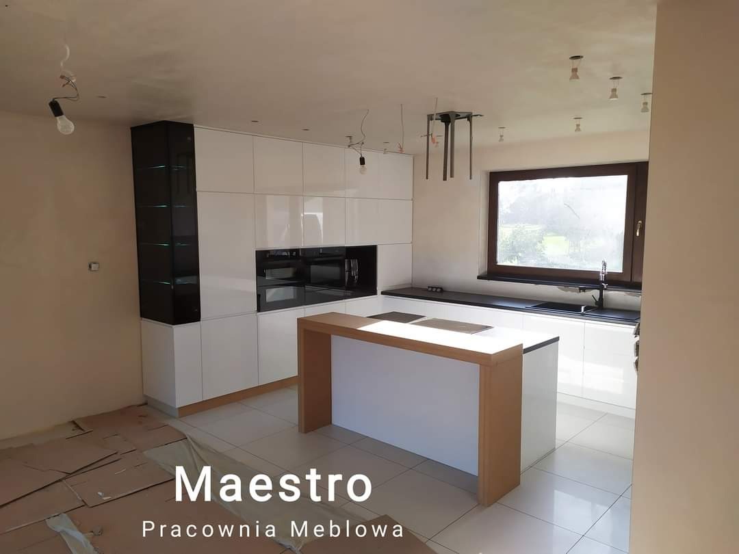 Nowoczesna kuchnia z białymi frontami i czarnym blatem, wyspa kuchenna z drewnianym blatem, widoczne niedokończone wykończenie sufitu i zabezpieczona podłoga.