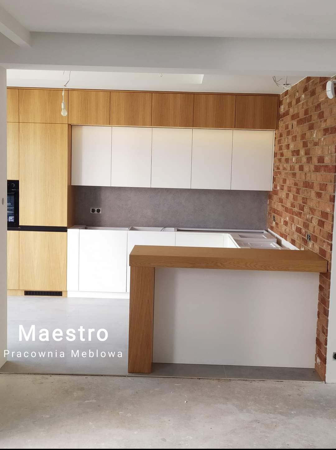 Jasna kuchnia z drewnianymi i białymi szafkami, surowa cegła na ścianie, widok na blat barowy z jasnego drewna, w trakcie montażu.