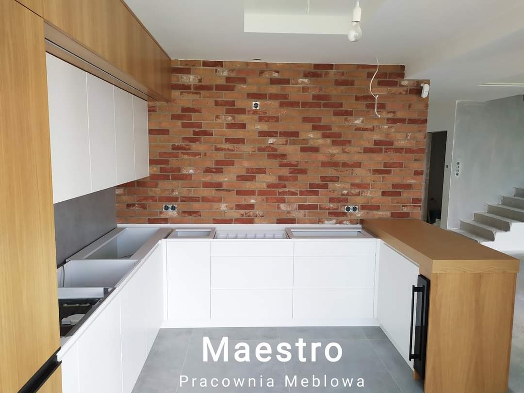 Montaż białej, nowoczesnej zabudowy kuchennej z drewnianym blatem i ceglaną ścianą w tle, widoczne niezabezpieczone kable i surowy stan wykończenia.