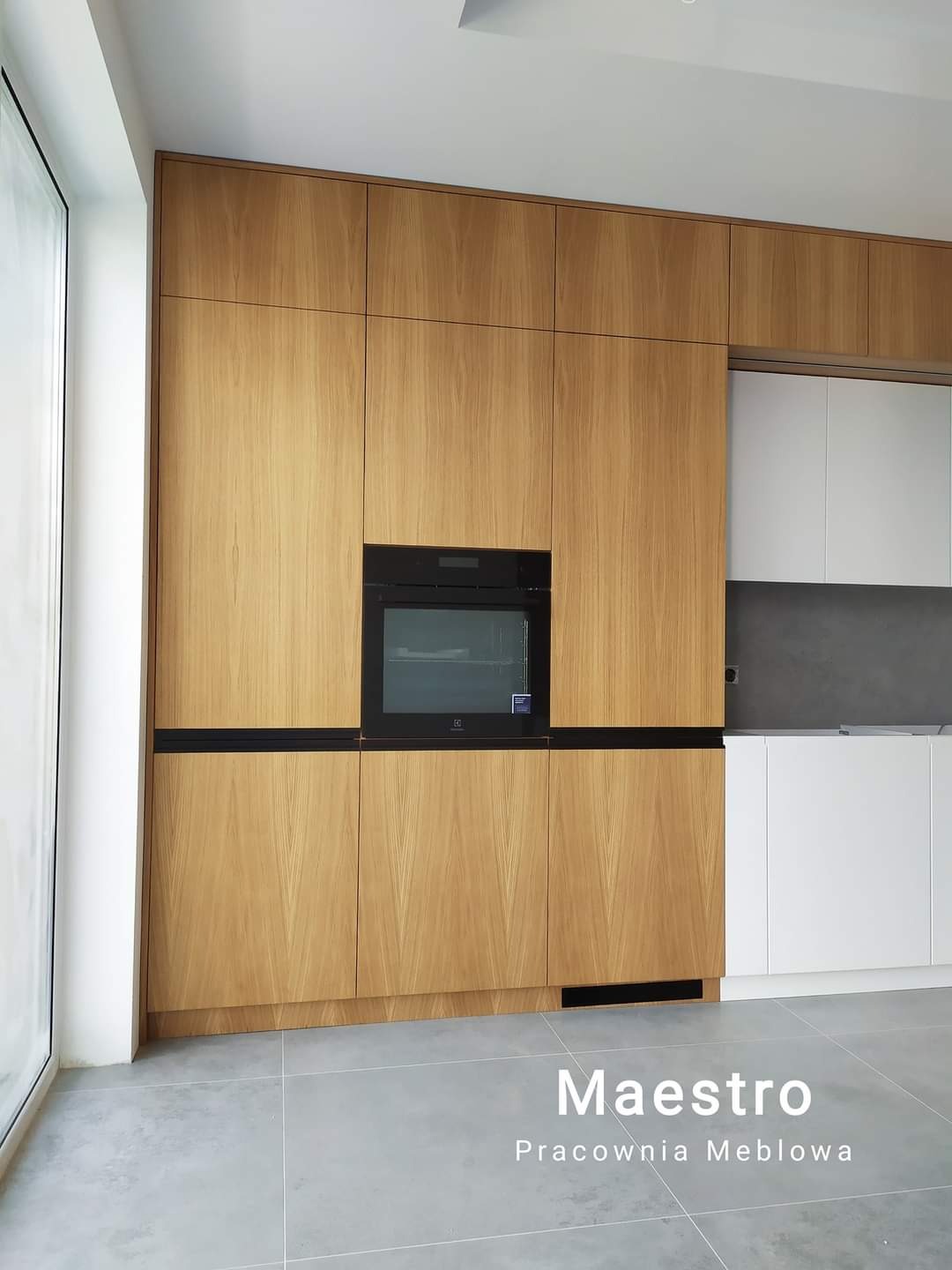 Nowoczesna zabudowa kuchenna z forniru dębowego z wbudowanym piekarnikiem i czarnymi akcentami, uzupełniona białymi szafkami i szarym blatem, widok z niskiego kąta.