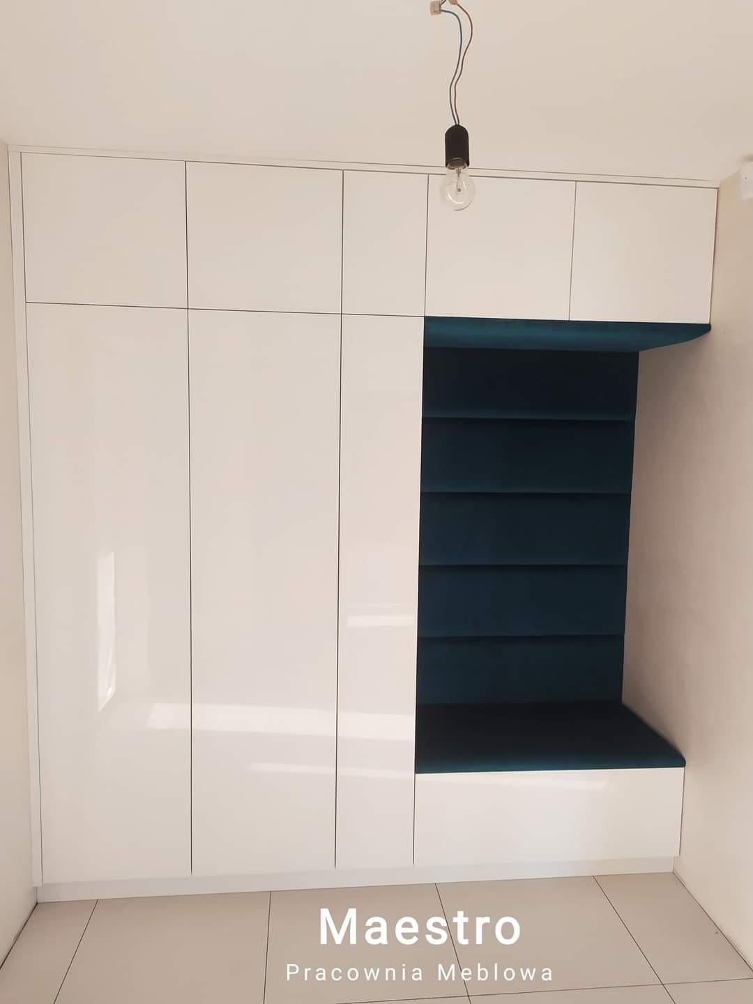 Biała szafa wnękowa z siedziskiem obitym granatowym aksamitem, oświetlona wiszącą żarówką bez klosza, na jasnej podłodze.