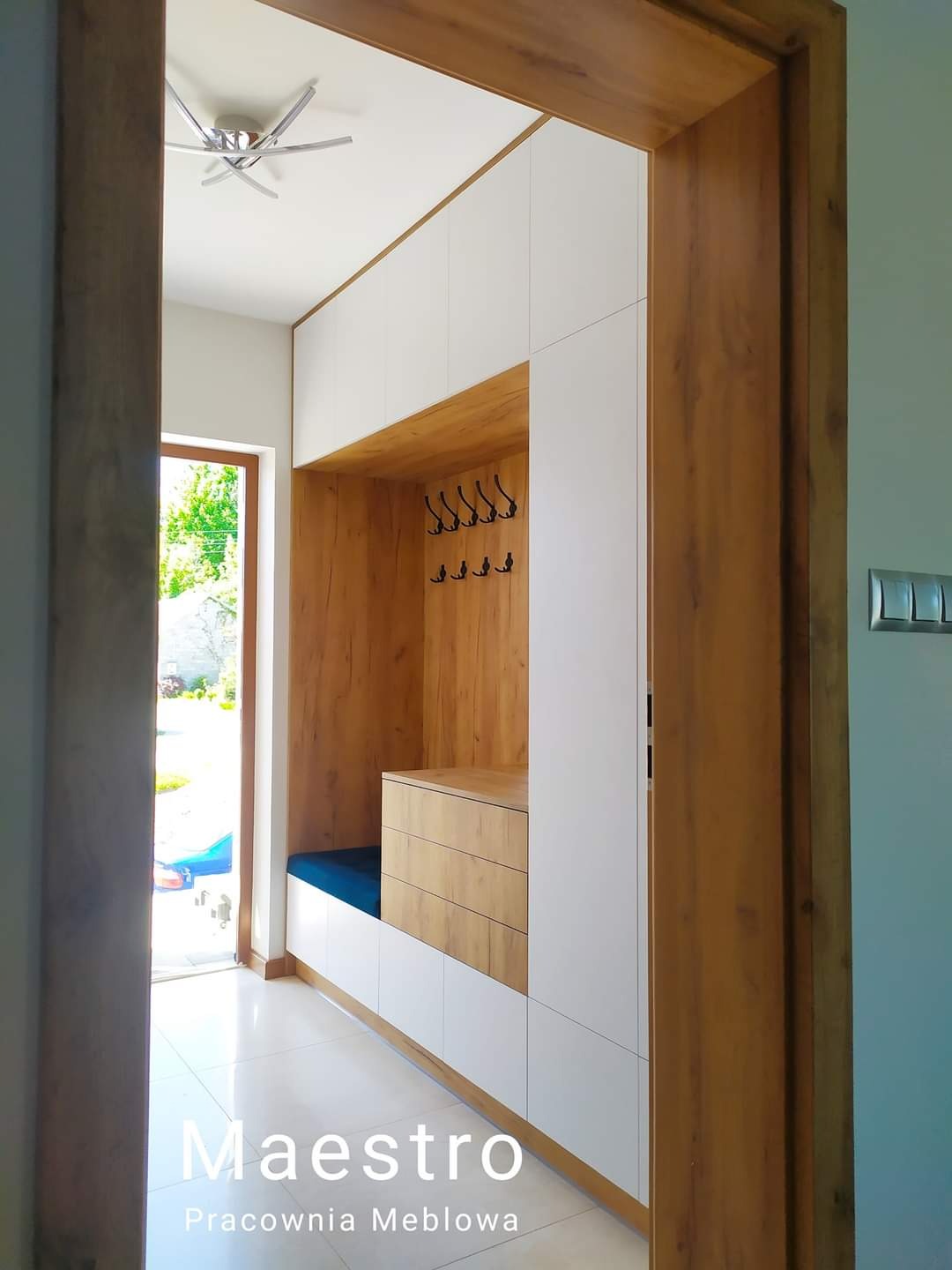 Wnęka garderobiana z siedziskiem i wieszakami, wykończona drewnem i bielą, widziana z perspektywy wejścia do pomieszczenia, z widokiem na zewnątrz przez okno.