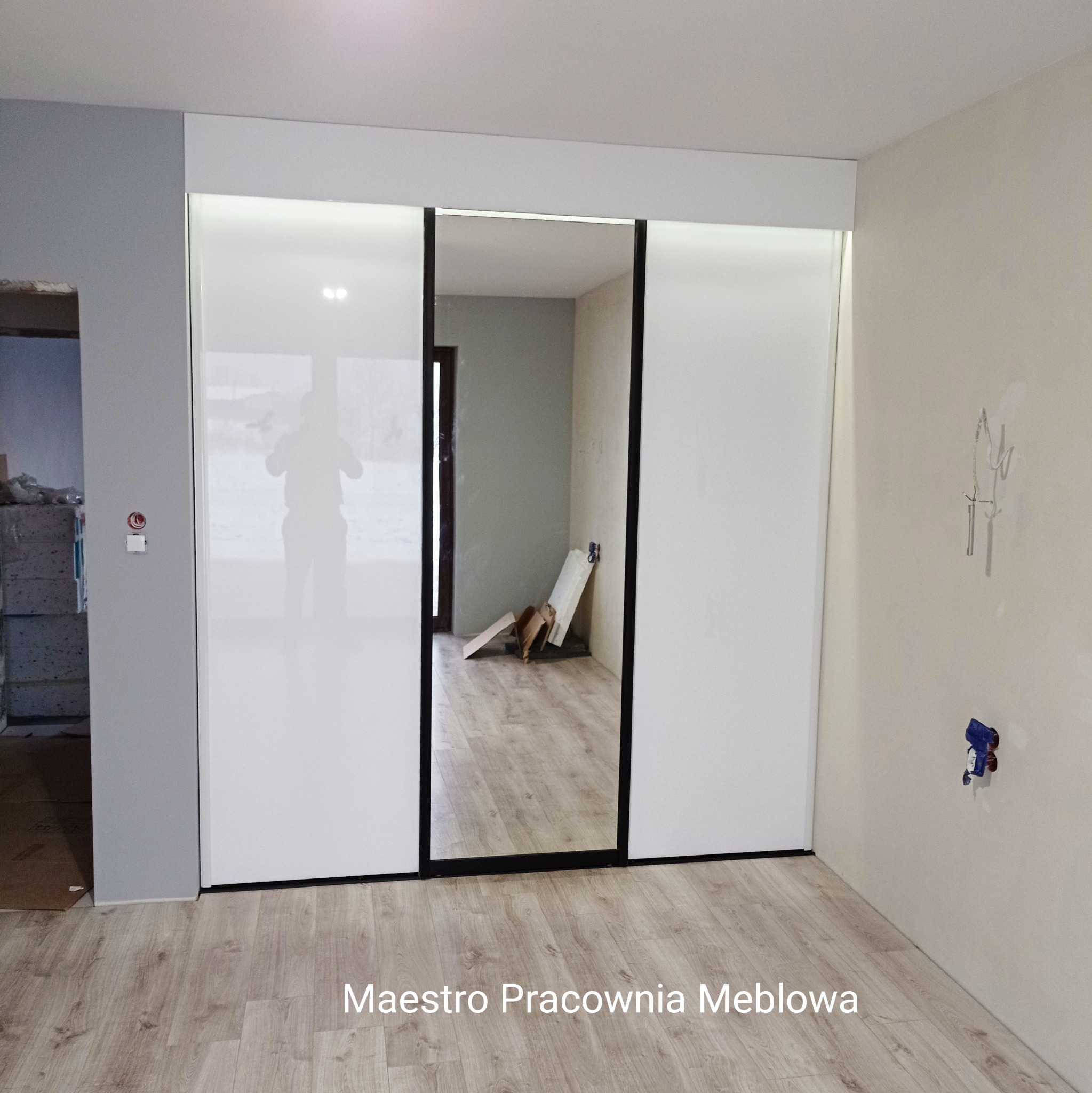 Biała szafa przesuwna z lustrem na środkowych drzwiach, widoczna w surowym wnętrzu z podłogą laminowaną i śladami po instalacjach na ścianie.