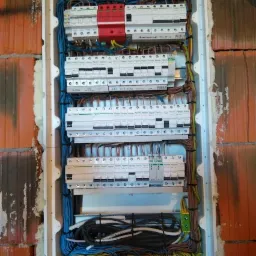 Rozdzielnica elektryczna w trakcie montażu, widoczne zabezpieczenia różnicowoprądowe, ograniczniki przepięć, przewody ułożone wewnątrz białej obudowy, tło z cegły i pianki montażowej.
