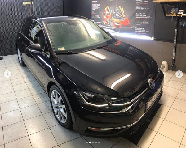Błyszczący, czarny Volkswagen Golf po detailingu, widoczny w studiu z plakatem o powłokach ochronnych na ścianie.