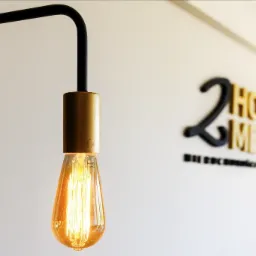 Edison bulb hanging from a black metal lamp stand with a brass fitting, against a white wall with a blurred company logo in the background.