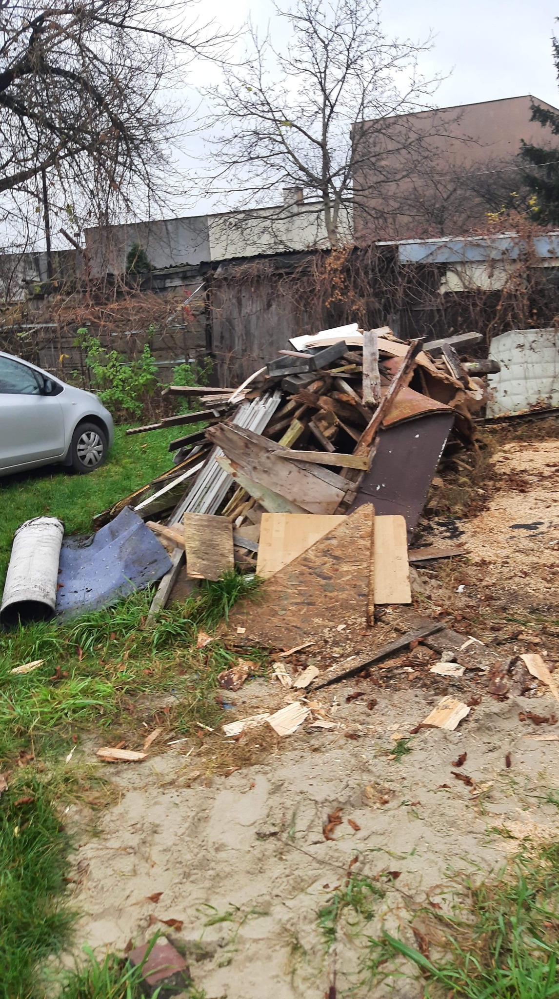 Stos drewnianych odpadów budowlanych, w tym deski, płyty i ramy okienne, leżący na trawie obok srebrnego samochodu osobowego w ogrodzie z drzewami bez liści w tle.