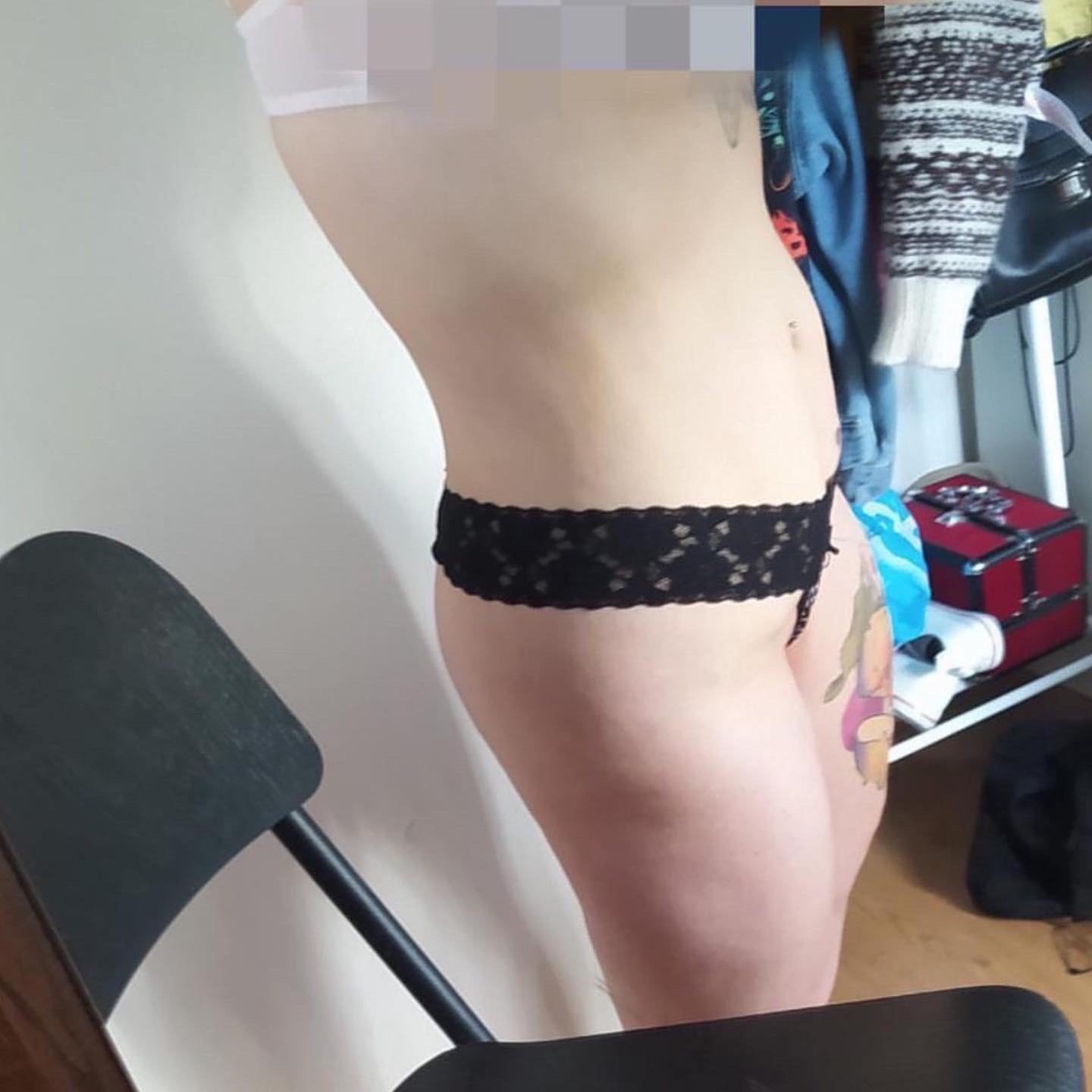 Profil sylwetki kobiety z tatuażem na udzie, w bieliźnie, na tle ściany i krzesła, widoczne ubrania i pudełko w tle.