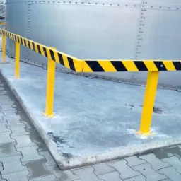 Żółto-czarna, metalowa barierka ochronna z odbojami, zamontowana na betonowym podłożu przy zbiorniku przemysłowym.