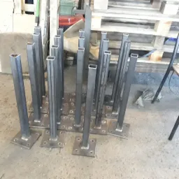Przygotowane do montażu, spawane, metalowe słupki o kwadratowym przekroju, z przyspawanymi podstawami z otworami na śruby, ułożone na podłodze warsztatu.