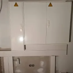 Biała szafka elektryczna z symbolem ostrzegawczym wysokiego napięcia zamontowana na ścianie w trakcie remontu, widoczne kable i puszki elektryczne.