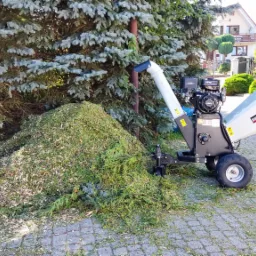 Stos świeżo rozdrobnionych gałęzi i igliwia obok profesjonalnego rębaka do gałęzi marki Rambo-HC15PRO, na tle zielonego drzewa iglastego i kostki brukowej.