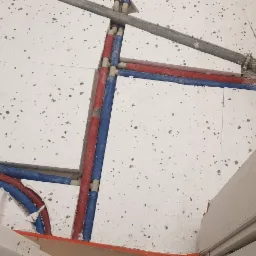 Podłoga w trakcie remontu z widocznymi rurami instalacji wodnej w kolorze czerwonym i niebieskim, częściowo ukrytymi w styropianie, oraz fragmentem ściany z widoczną instalacją.