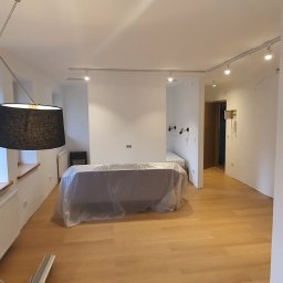 ROT Mayer - Jasne, przestronne wnętrze mieszkania z parkietem, białymi ścianami, osłoniętym meblem i nowoczesnym oświetleniem sufitowym oraz lampami z czarnymi abażurami, prawdopodobnie świeżo po malowaniu.