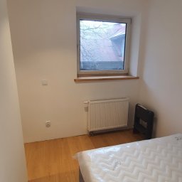 ROT Mayer - Pokój połóżony na parkiecie z białymi ścianami, grzejnikiem pod oknem i nowym materacem z folii na łóżku.