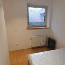 Pokój połóżony na parkiecie z białymi ścianami, grzejnikiem pod oknem i nowym materacem z folii na łóżku.