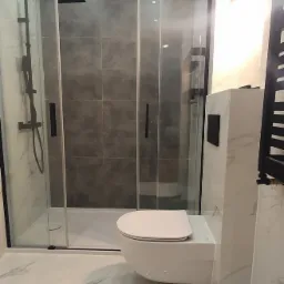 Nowoczesna łazienka po remoncie: białe marmurowe płytki, podwieszana toaleta z czarnym przyciskiem, kabina prysznicowa z czarną ramą i deszczownicą