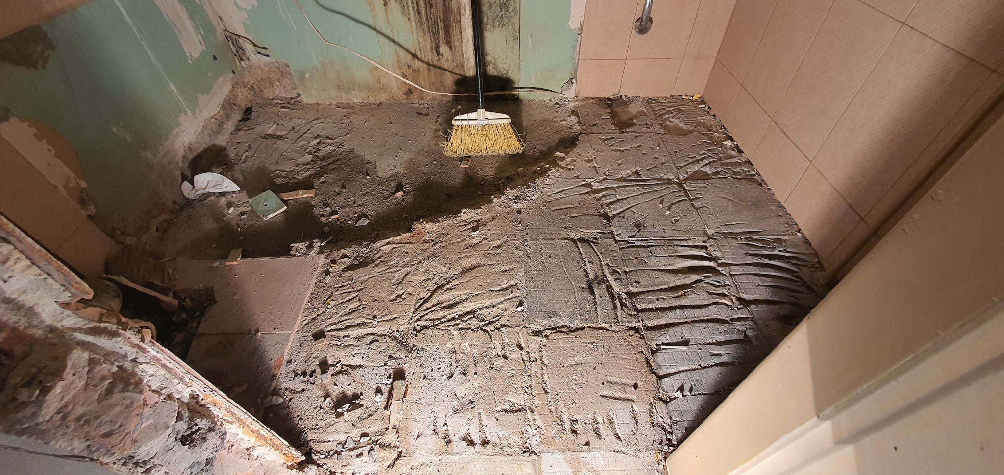 Demontaż płytek podłogowych w łazience, widoczne pozostałości kleju i zaprawy, ściany częściowo bez okładziny, stojąca miotła