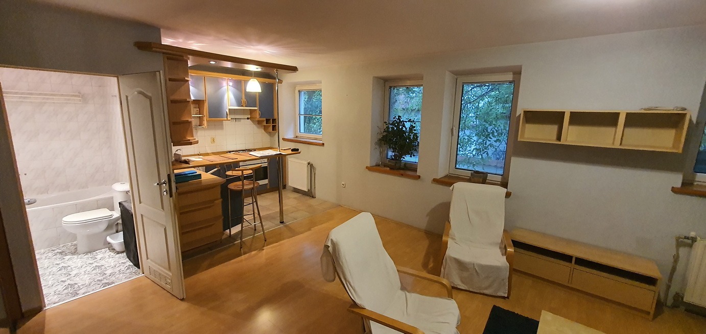 Widok na otwartą przestrzeń mieszkania po remoncie, obejmującą łazienkę z białą armaturą, kuchnię z drewnianymi szafkami i barem z hokerami, oraz część wypoczynkową z dwoma fotelami przykrytymi...