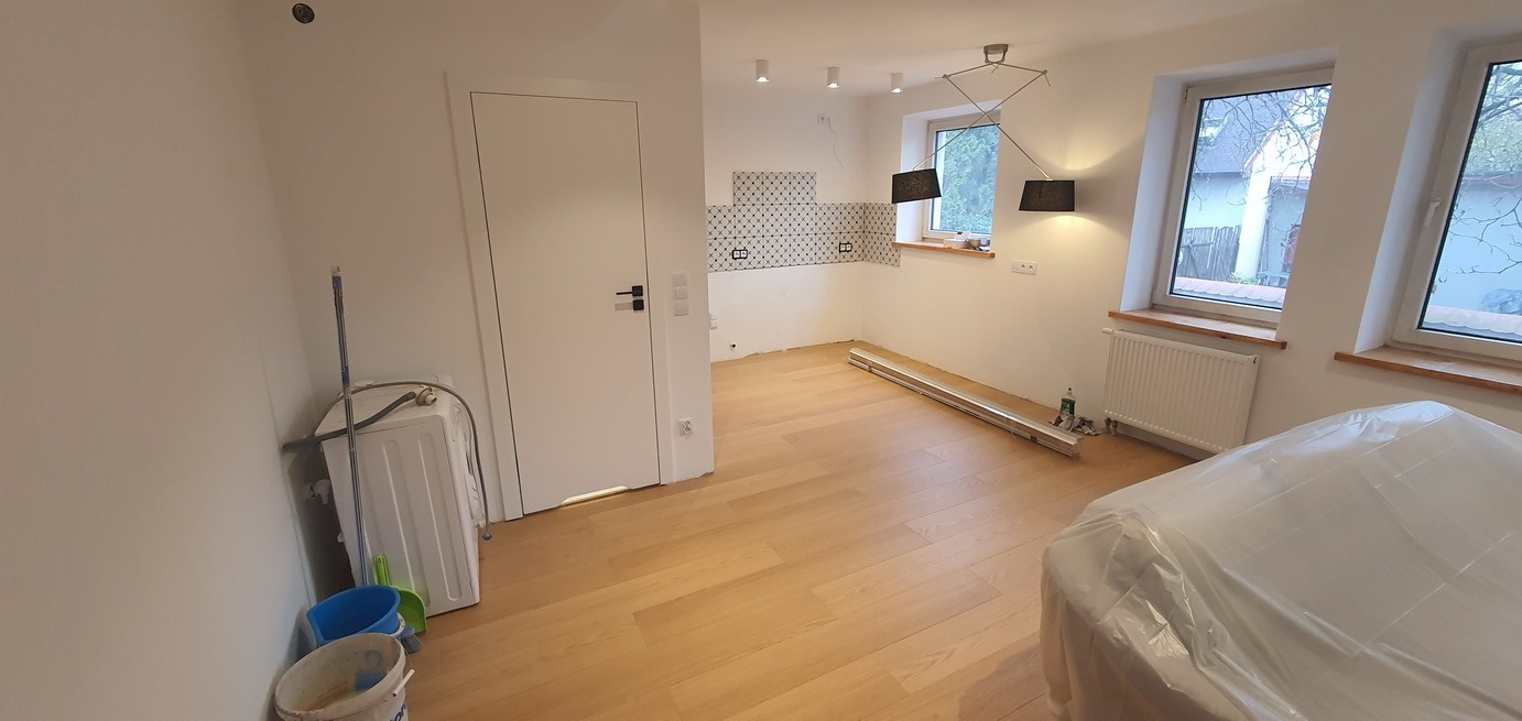 Wnętrze w trakcie remontu: świeżo położony parkiet, częściowo ułożone płytki na ścianie, zakryte meble folią, widoczne narzędzia i wiadro, pralka w rogu pomieszczenia.