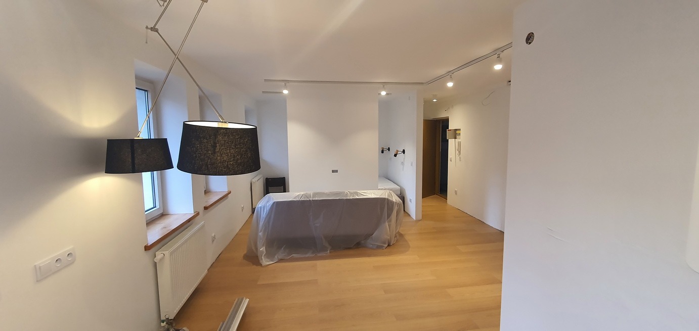 Jasne, przestronne wnętrze mieszkania z parkietem, białymi ścianami, osłoniętym meblem i nowoczesnym oświetleniem sufitowym oraz lampami z czarnymi abażurami, prawdopodobnie świeżo po malowaniu.