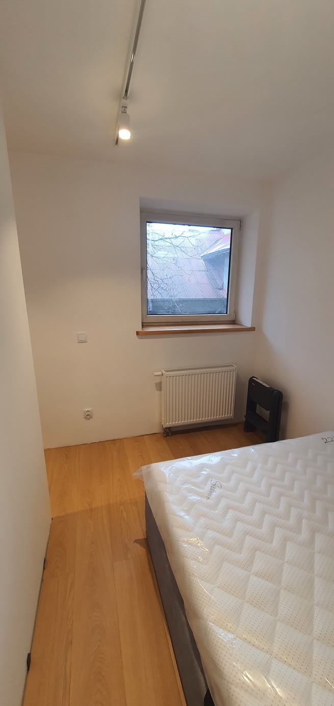 Pokój połóżony na parkiecie z białymi ścianami, grzejnikiem pod oknem i nowym materacem z folii na łóżku.