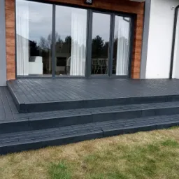Szary taras z kostki brukowej przed domem z dużymi przesuwnymi drzwiami, widok z poziomu trawnika.