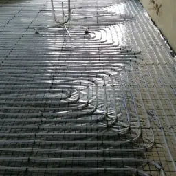 Rozłożone rury ogrzewania podłogowego na folii z nadrukiem, widoczne mocowania rur, w tle okno i surowa ściana.